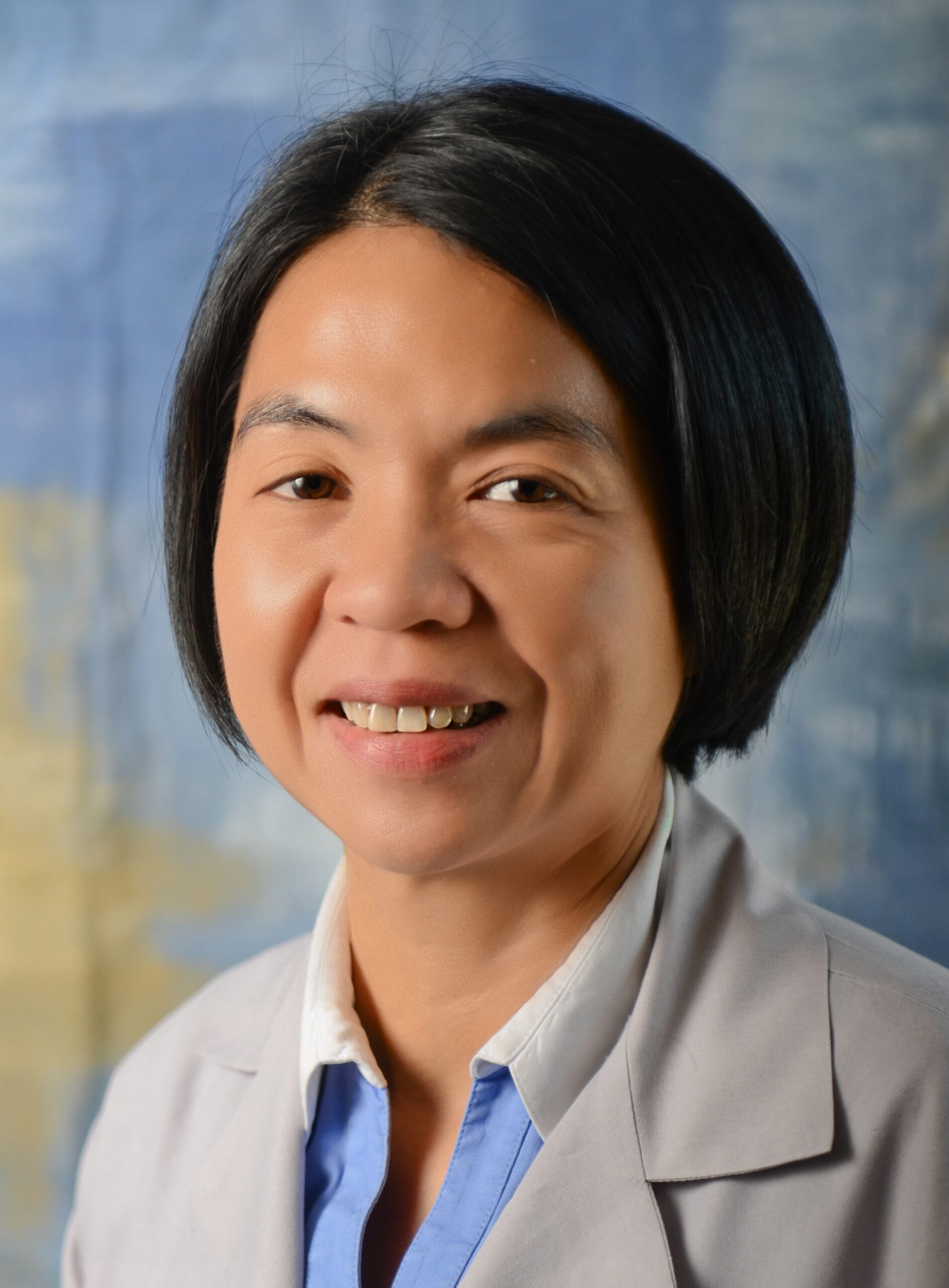 Dr. Lorraine Y. Bangayan is pictured with short black hair smiles wearing a light gray blazer over a blue shirt. The background is a softly blurred mix of blue and beige colors.